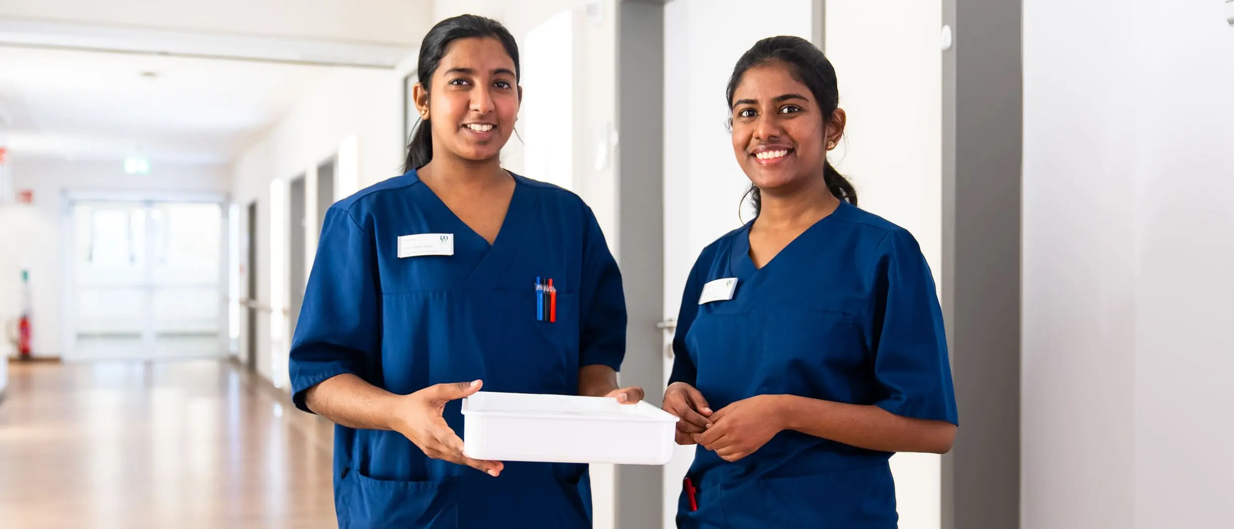 Zwei Krankenschwestern in blauen Uniformen stehen im Krankenhausflur und halten eine wei&szlig;e Box.
