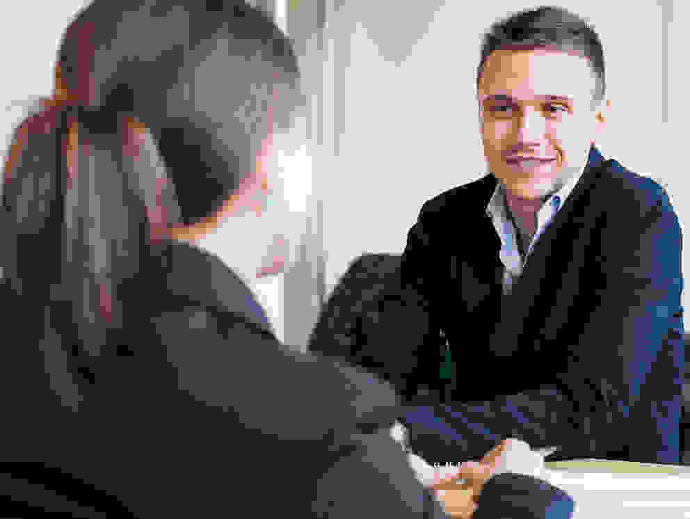 Zwei Personen in Anzügen sitzen an einem Tisch und führen ein Gespräch. Im Hintergrund sind weitere Personen an anderen Tischen zu sehen.