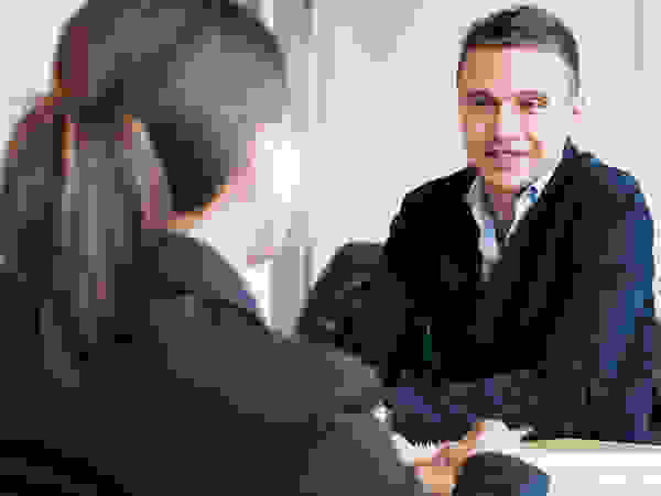 Zwei Personen in Anzügen sitzen an einem Tisch und führen ein Gespräch. Im Hintergrund sind weitere Personen an anderen Tischen zu sehen.