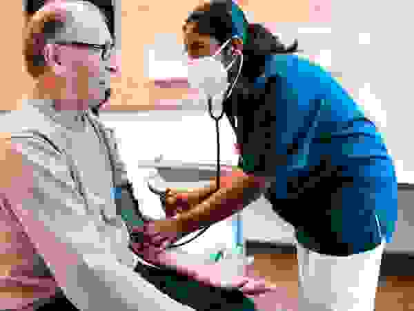 Ein älterer Mann sitzt auf einem Bett, während eine Krankenschwester mit einer Maske seinen Blutdruck misst.