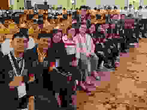 Gruppenfoto von Teilnehmern eines Events in einem Saal, in dem viele Menschen mit orangefarbenen Jacken sitzen. Plakate mit Bildern von Führungspersönlichkeiten sind im Hintergrund sichtbar.
