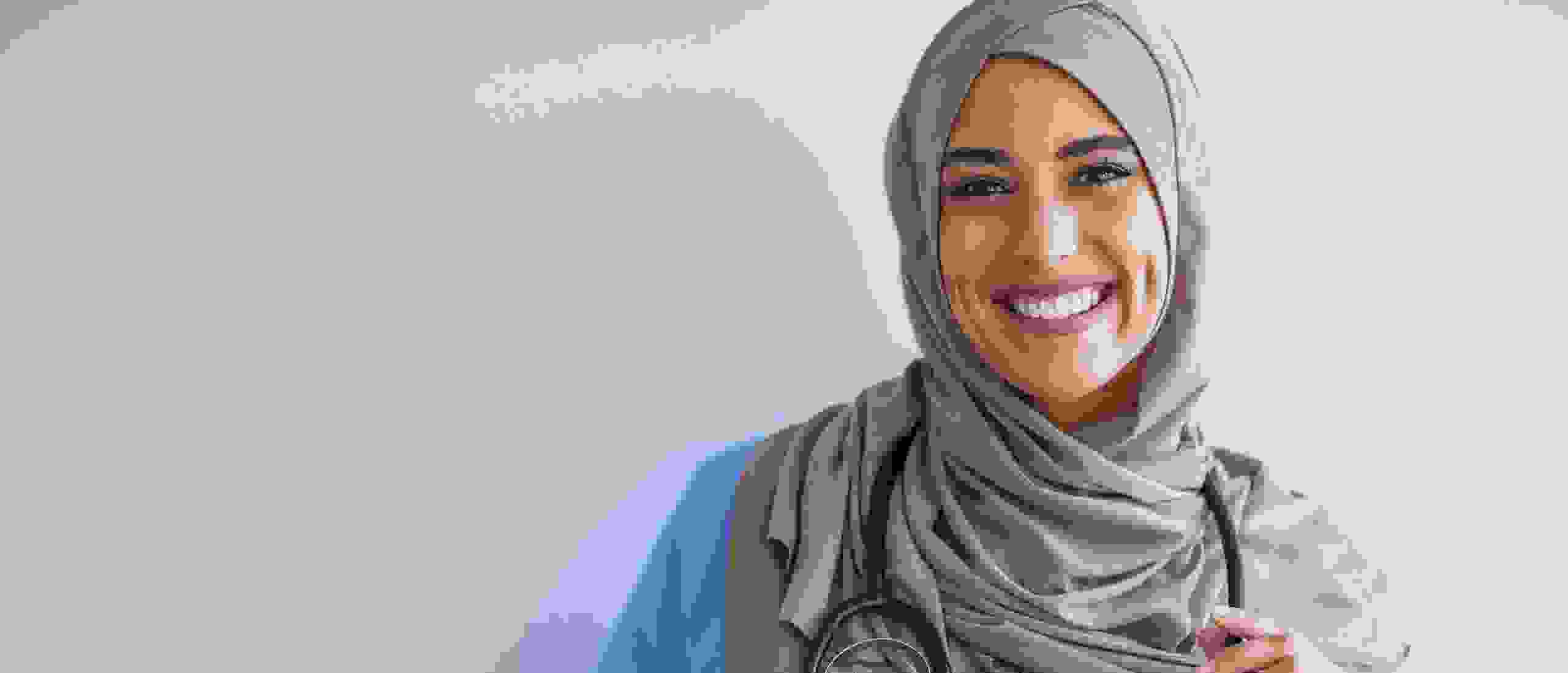 Eine lächelnde medizinische Fachkraft in blauer Kleidung und einem Hijab, die ein Stethoskop hält.