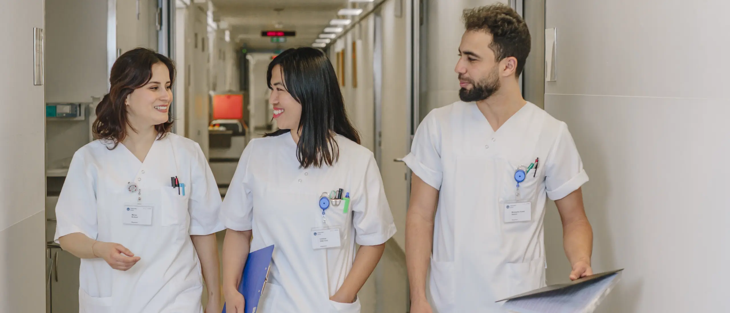 Drei medizinische Fachkräfte in weißen Arbeitskleidungen, die lächelnd einen Flur im Krankenhaus entlanggehen, dabei Notizen und Dokumente halten.