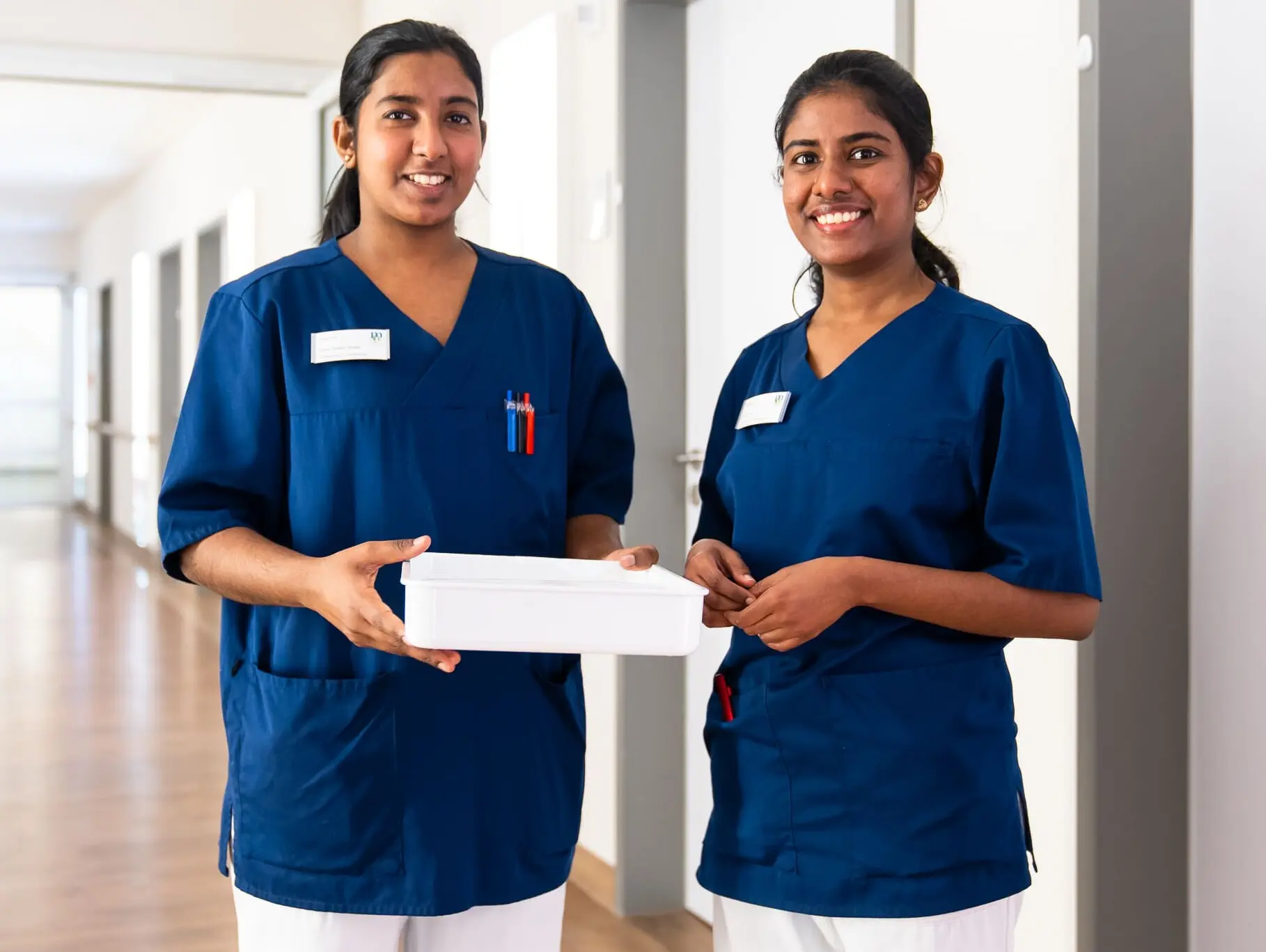 Zwei Krankenschwestern in blauen Uniformen stehen im Krankenhausflur und halten eine wei&szlig;e Box.