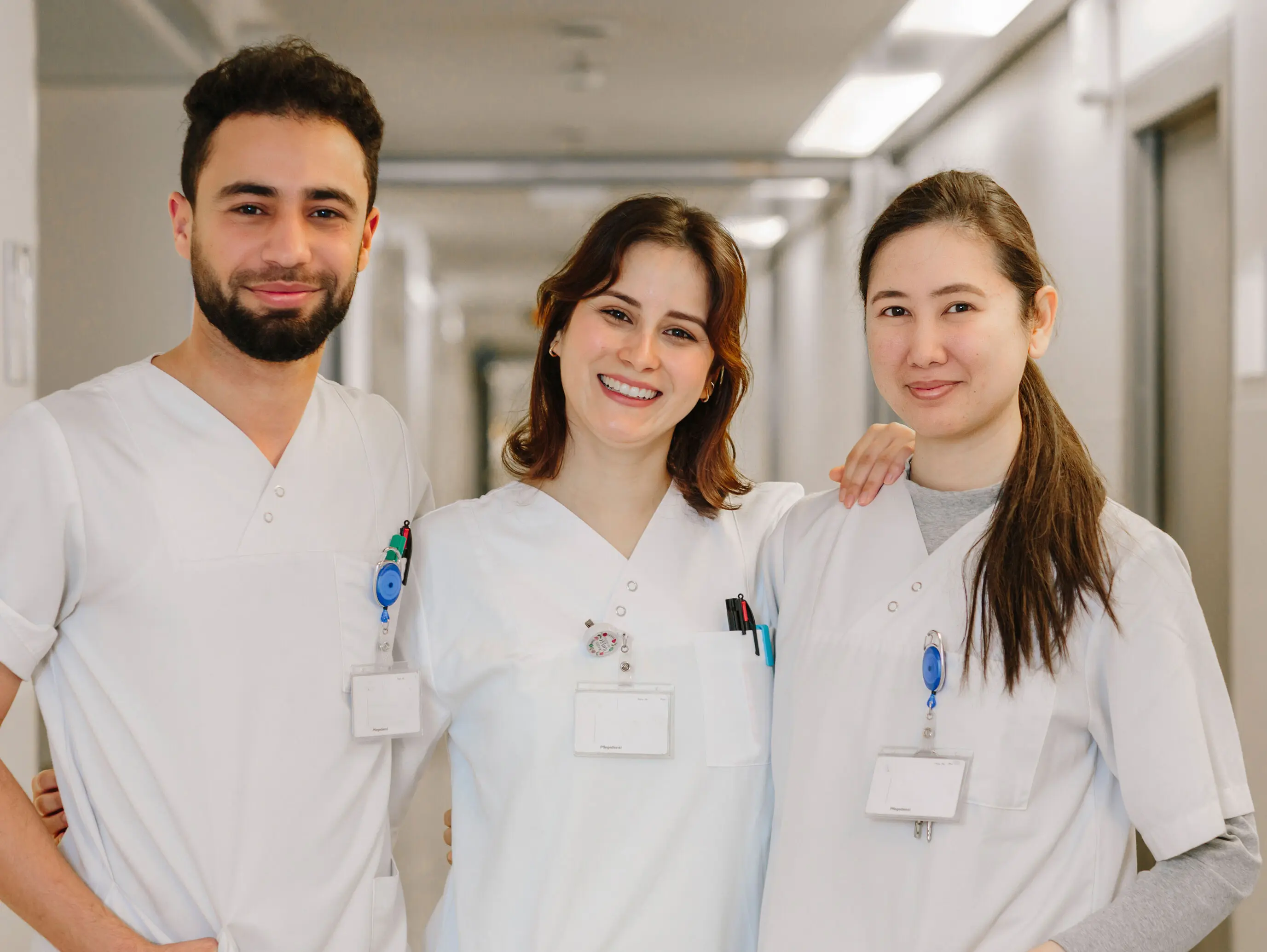 Drei fröhliche Pflegekräfte in weißen Arbeitsuniformen stehen in einem Krankenhausflur und lächeln in die Kamera.