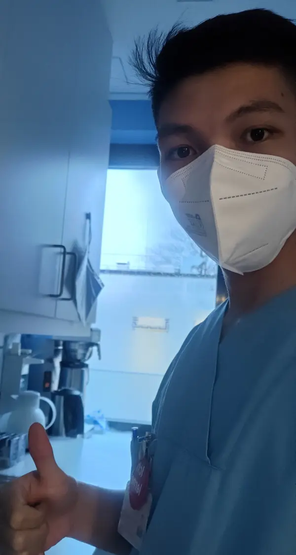 Junger Mann in blauer Arbeitskleidung und einer medizinischen Maske, der einen Daumen nach oben zeigt, steht in einer hellen Klinik-Umgebung.