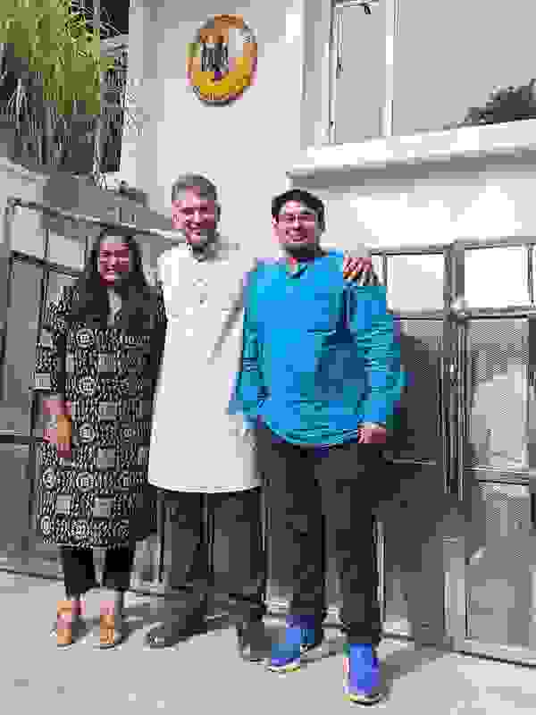 Drei Personen stehen vor einem Gebäude mit einem Wappen. Eine Frau in einem schwarzen Kleid, ein Mann in einem weißen Kurta und ein Mann in einem blauen Hemd posieren lächelnd.