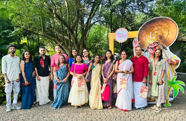 Eine Gruppe von Menschen in traditioneller indischer Kleidung posiert vor einem gr&uuml;nen Hintergrund und einem farbenfrohen, geschnitzten Charakter.