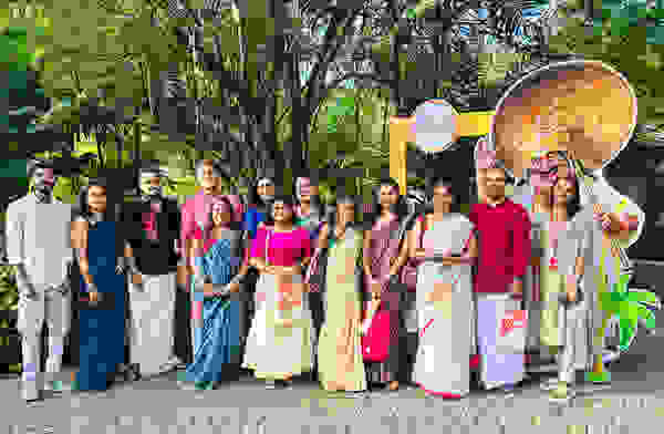 Eine Gruppe von Menschen in traditioneller indischer Kleidung posiert vor einem grünen Hintergrund und einem farbenfrohen, geschnitzten Charakter.