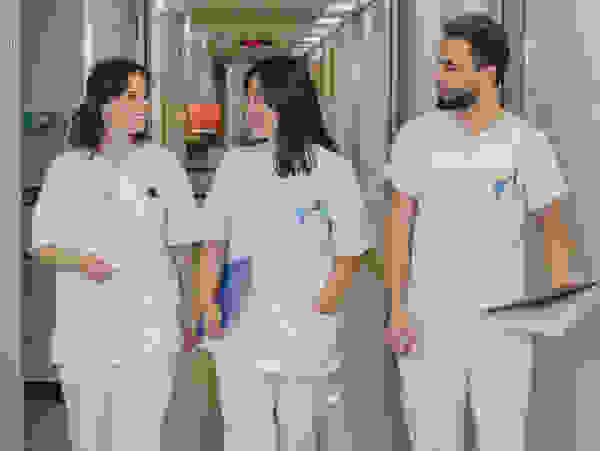 Drei Krankenpflegekräfte in weißen Arbeitsuniformen gehen lächelnd einen Flur im Krankenhaus entlang.