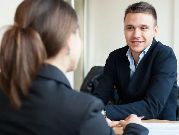 Two people in suits are sitting at a table having a conversation. Other people can be seen at other tables in the background.