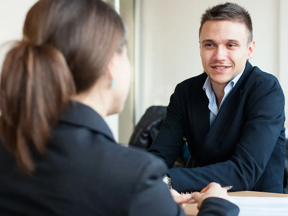 Two people in suits are sitting at a table having a conversation. Other people can be seen at other tables in the background.