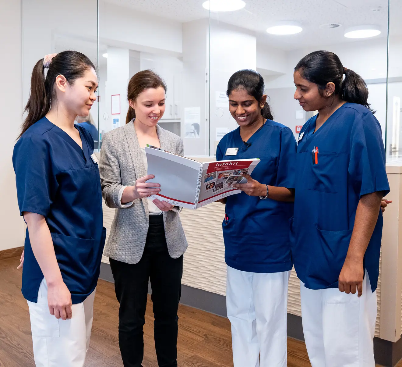 Four people in a modern medical environment are looking at a book together. Three are wearing blue work clothes, while one person is dressed in a gray blazer.