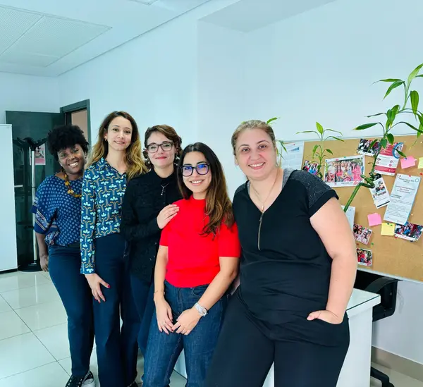 Five women are standing in an office, smiling and posing together. Behind them is a wall board with notes and plants.