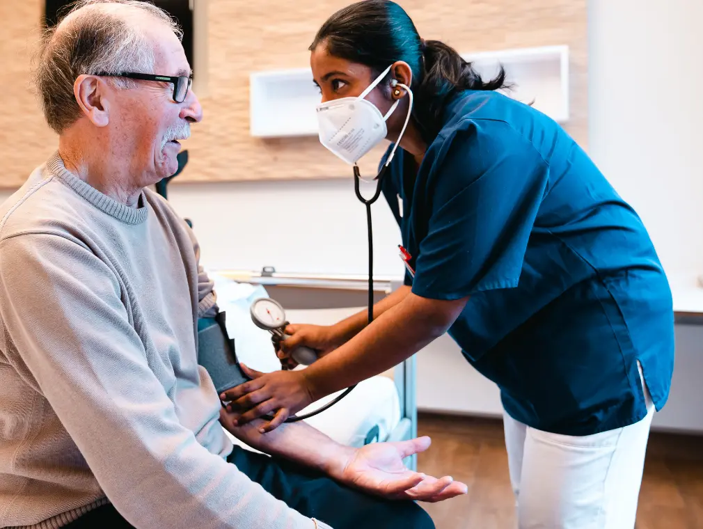An elderly man sits on a bed while a nurse with a mask measures his blood pressure.