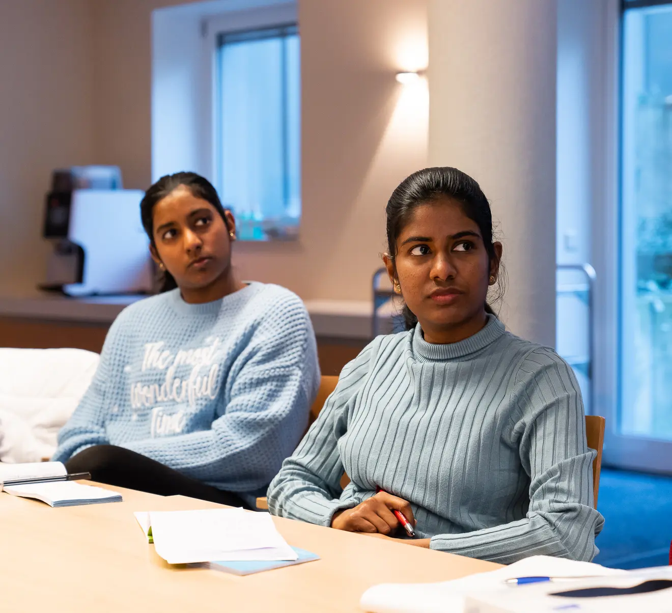 Two women are sitting at a table in a meeting room. One woman is wearing a light blue, structured sweater, the other a blue turtleneck sweater, both are listening attentively. A man can also be seen in the picture, but his focus is not on the camera.
