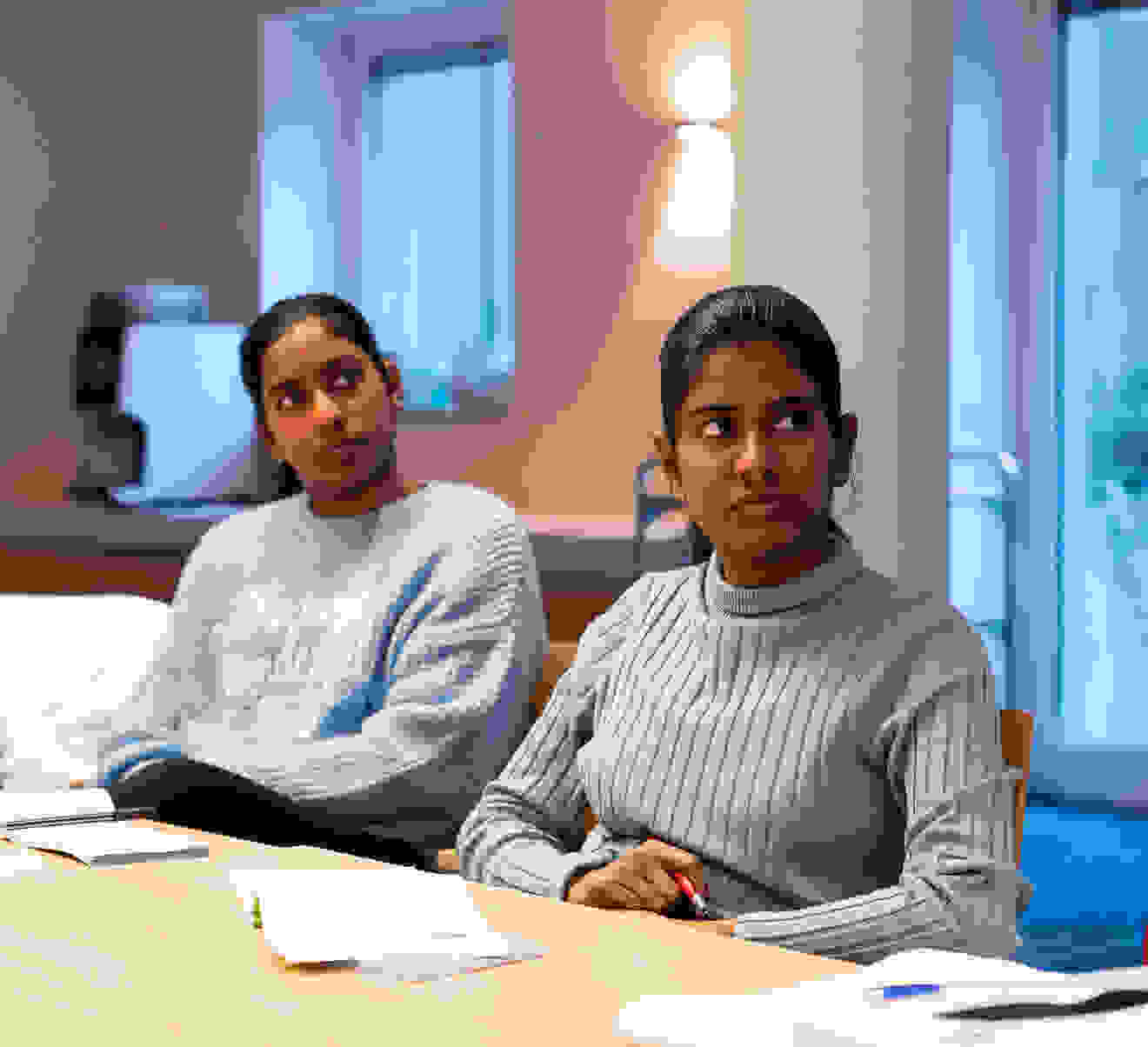 Two women are sitting at a table in a meeting room. One woman is wearing a light blue, structured sweater, the other a blue turtleneck sweater, both are listening attentively. A man can also be seen in the picture, but his focus is not on the camera.