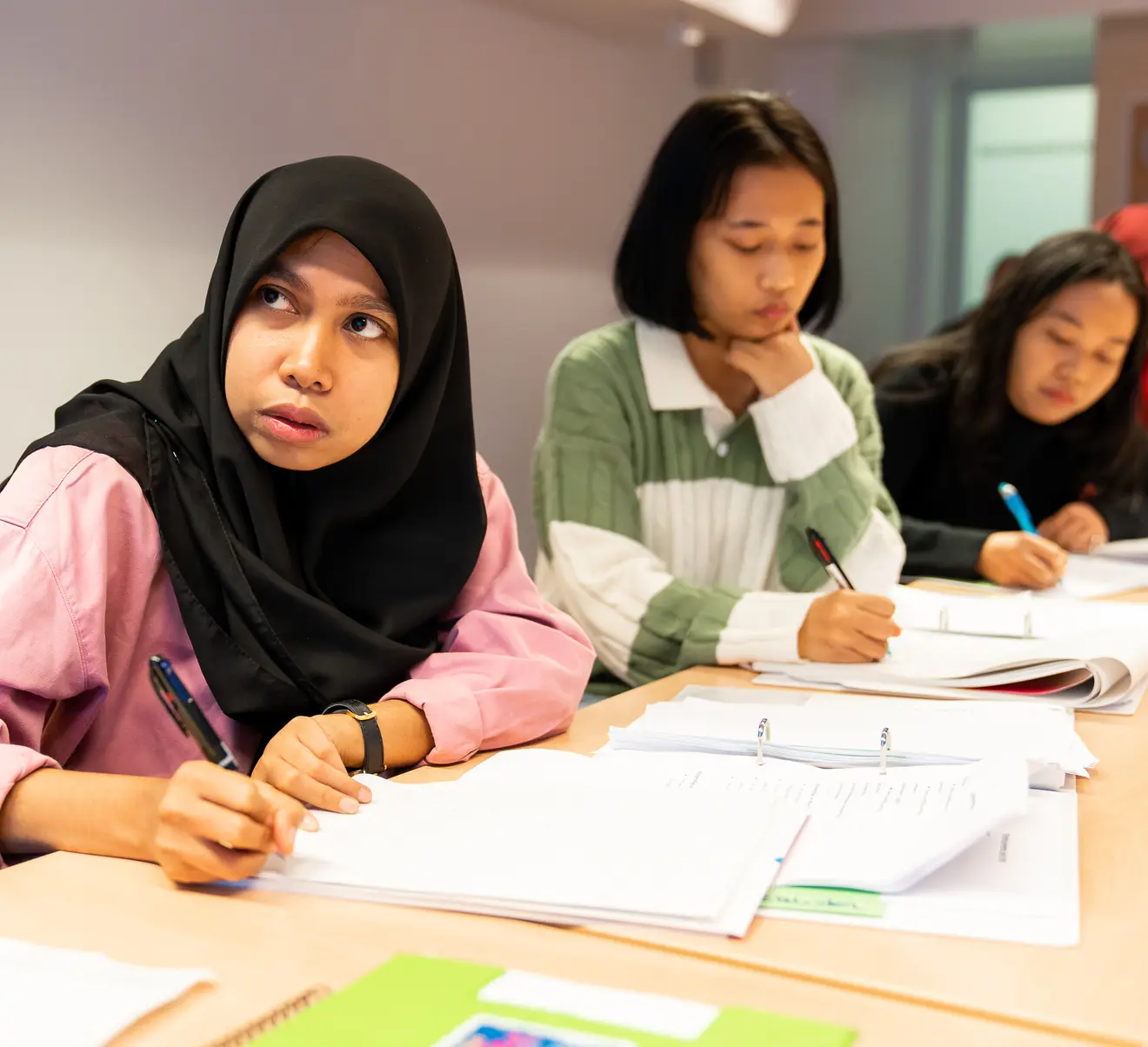 A group of young women sit at a table, focused on their notes and writing as they take part in a class.
