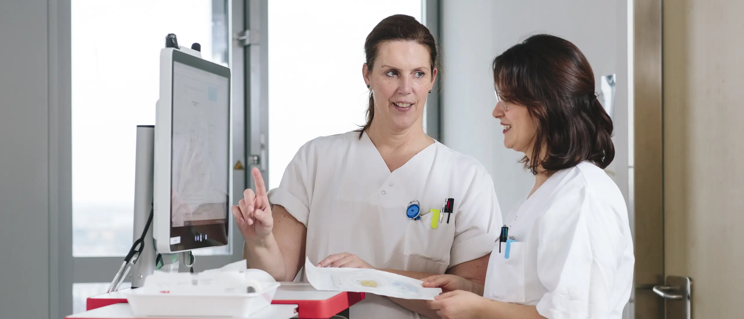 Two nurses in white uniforms discussing patient data at a computer workstation in a modern hospital.