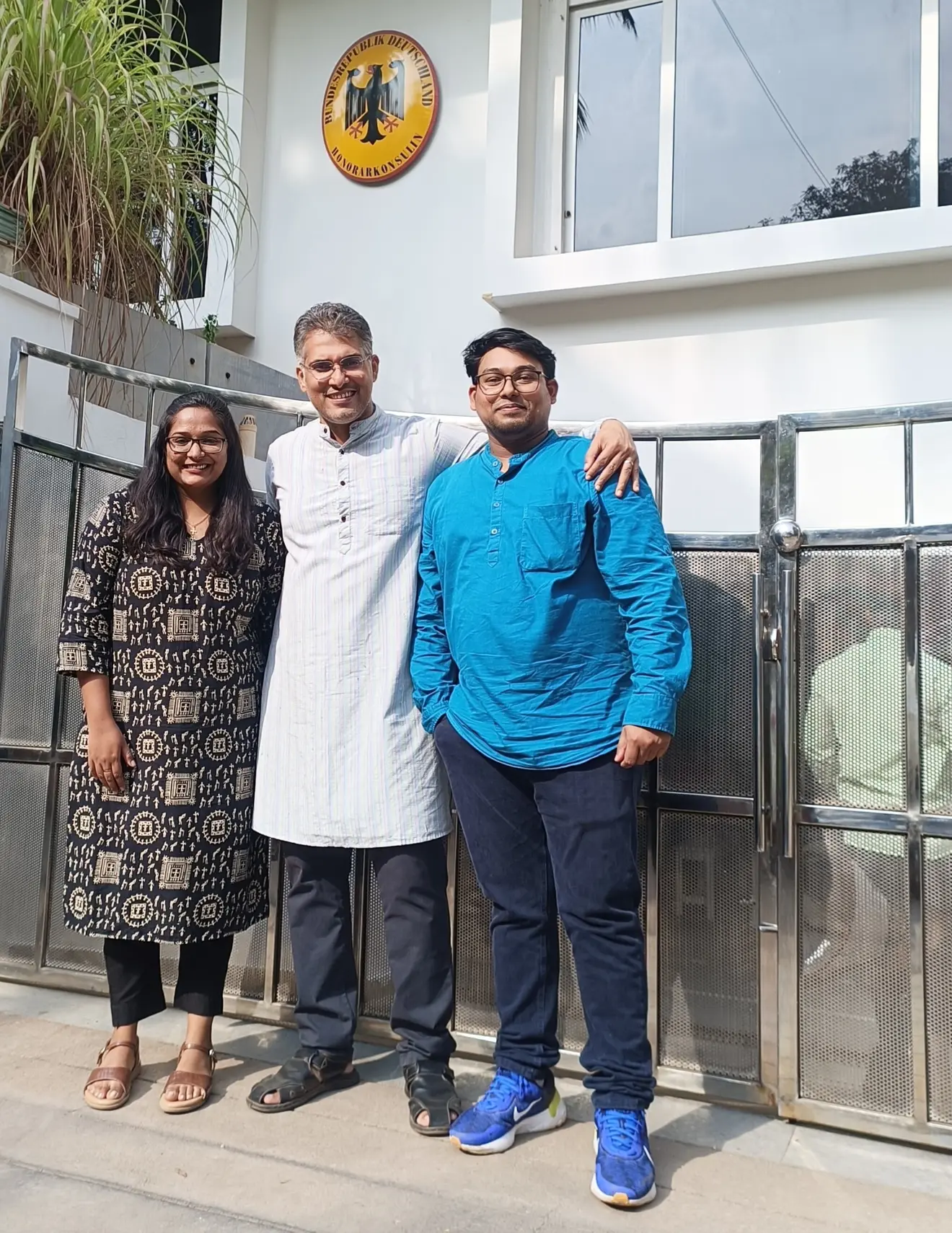 Three people stand in front of a building with a coat of arms. A woman in a black dress, a man in a white kurta and a man in a blue shirt pose smiling.