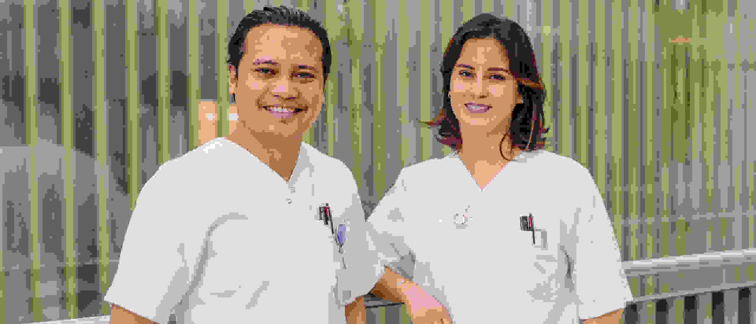 Two smiling healthcare professionals in white coats stand in front of a modern glass wall with vertical stripes.