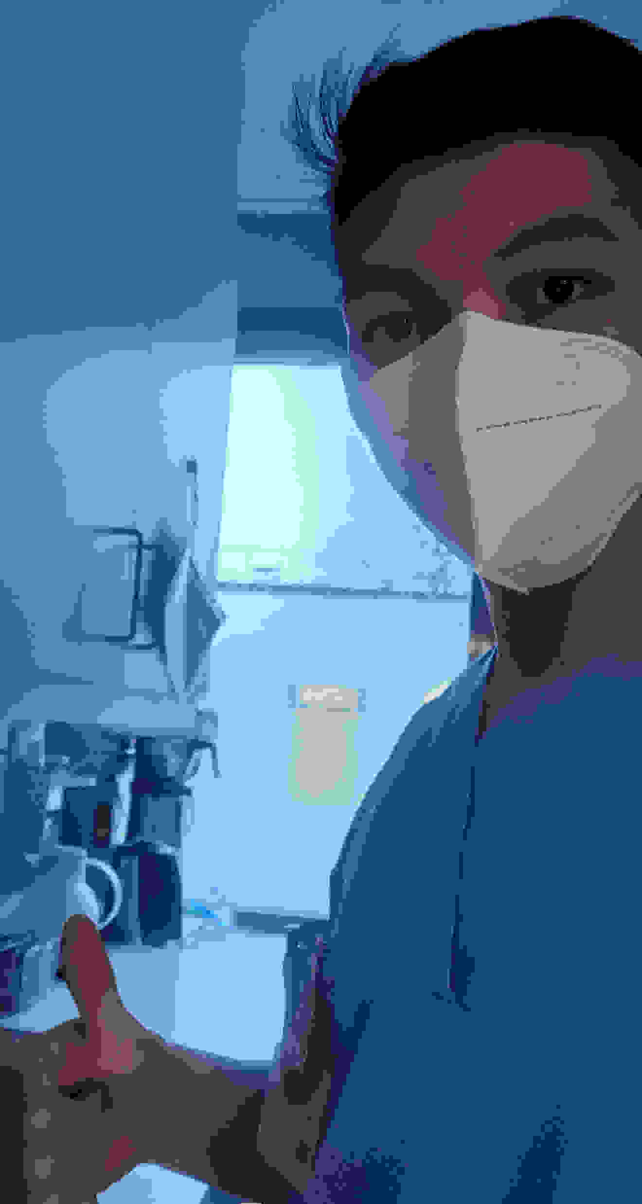 Young man in blue work clothes and a medical mask, giving a thumbs up, stands in a bright clinic environment.