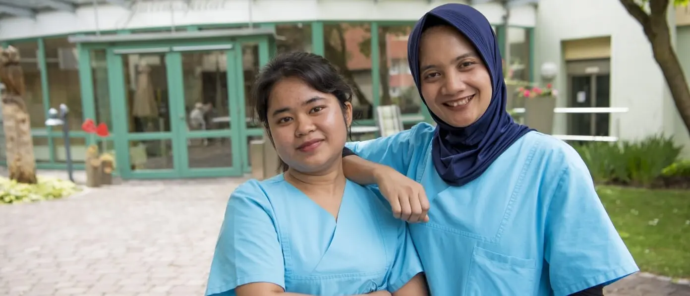 Two women in blue nursing uniforms smile in front of a building.