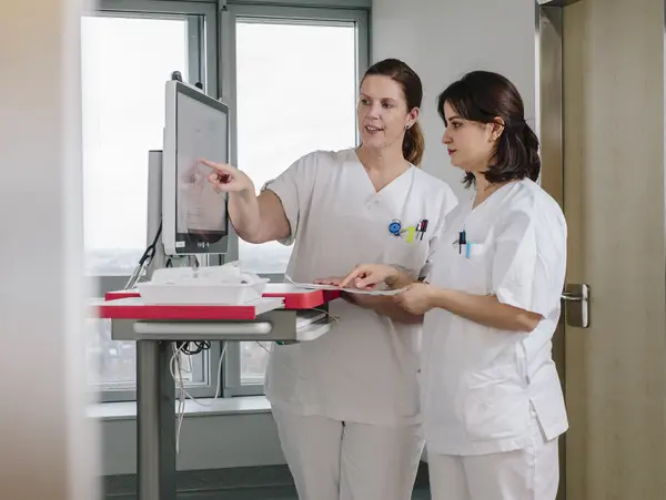 Two nurses in white coats look together at a screen in a hospital corridor.