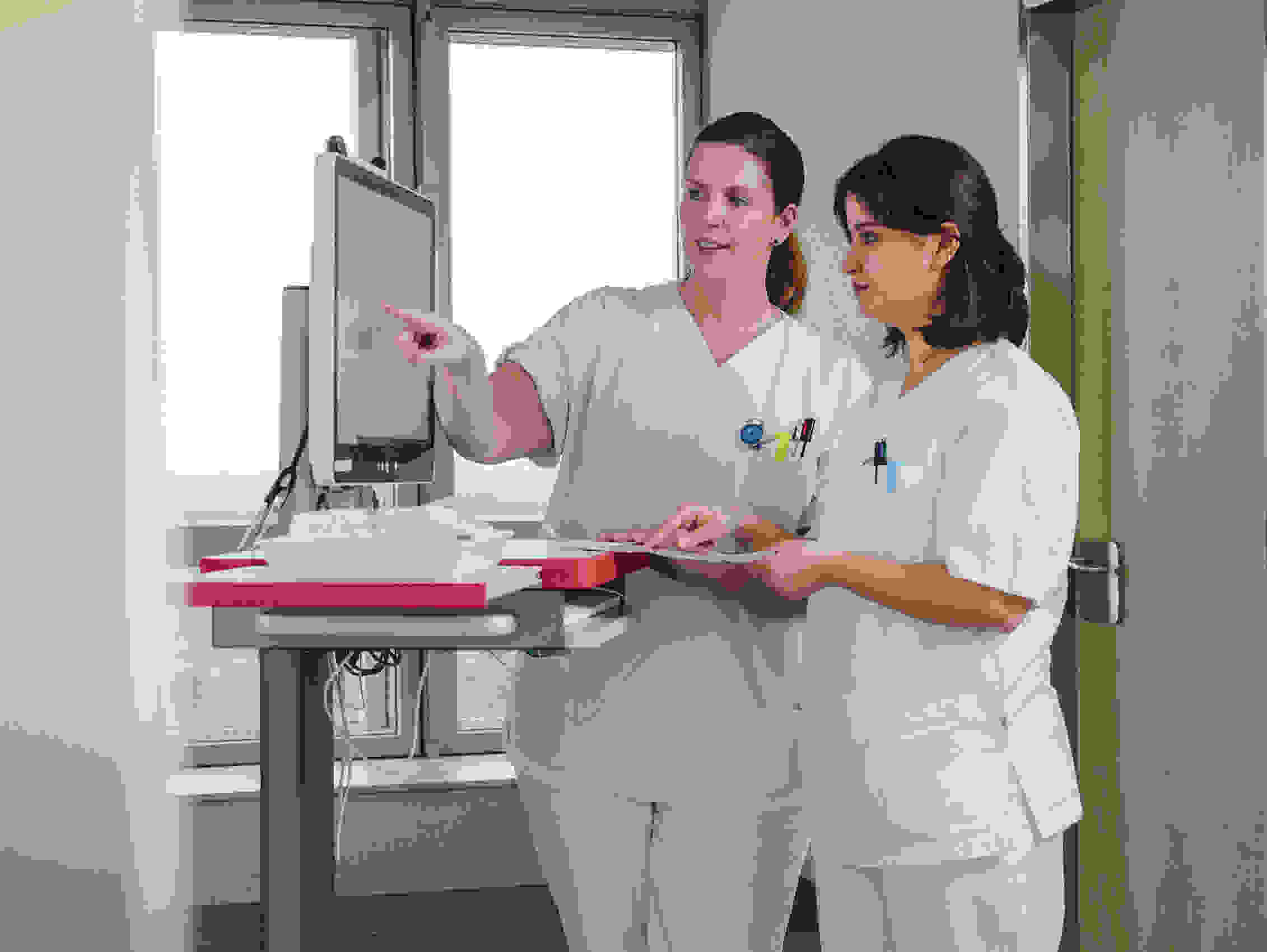 Two nurses in white coats look together at a screen in a hospital corridor.