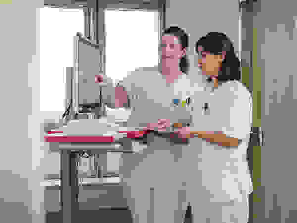 Two nurses in white coats look together at a screen in a hospital corridor.