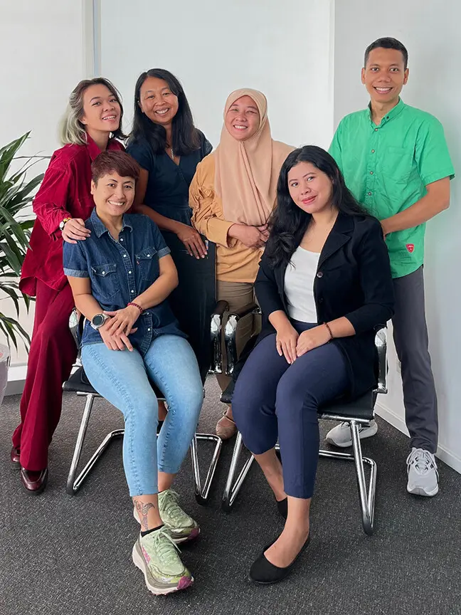Group of six people in a modern office, posing for a photo. They stand and sit together, surrounded by an arrangement of plants.