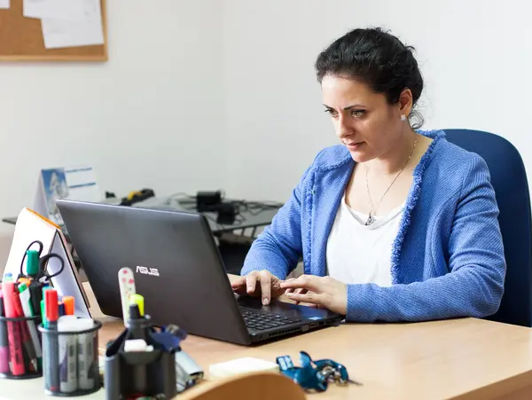 A woman sits at a desk and works concentrated on a laptop. There are various office supplies on the table and a map on the wall.