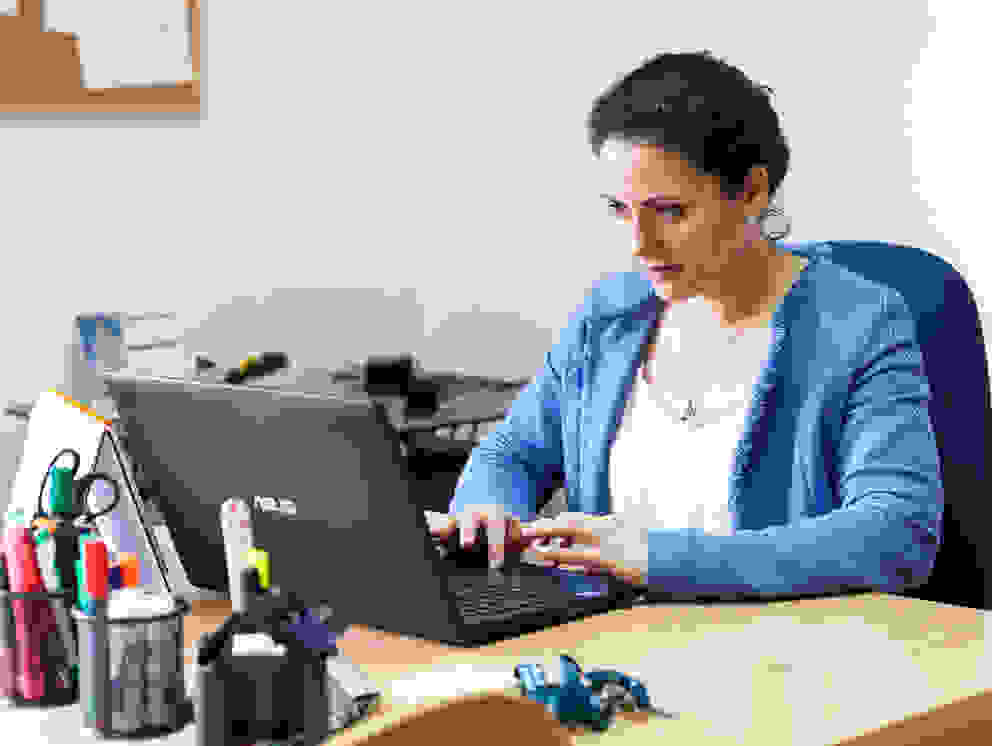 A woman sits at a desk and works concentrated on a laptop. There are various office supplies on the table and a map on the wall.