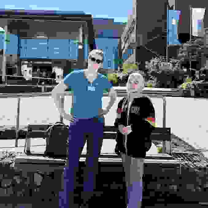 Two people are standing in front of a modern building. One woman is wearing a blue shirt and sunglasses, the other a black sweatshirt with colorful stripes. Both are smiling slightly.