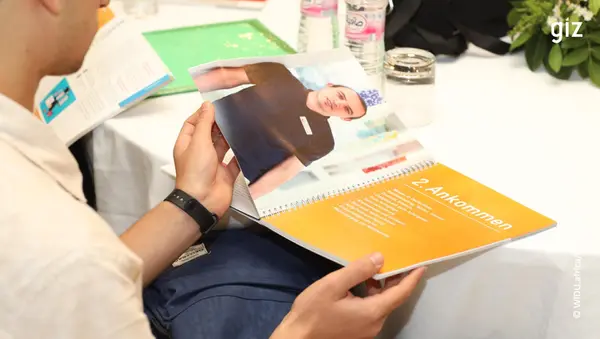 A person is holding an open booklet with a picture of a man in a dark shirt. The title on one page reads “2nd arrival” in German. Water bottles and leaves can be seen in the background.