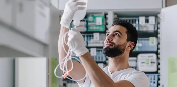 A healthcare worker holds an infusion bottle in his hand and looks at it attentively. Medication and medical equipment are visible in the background.