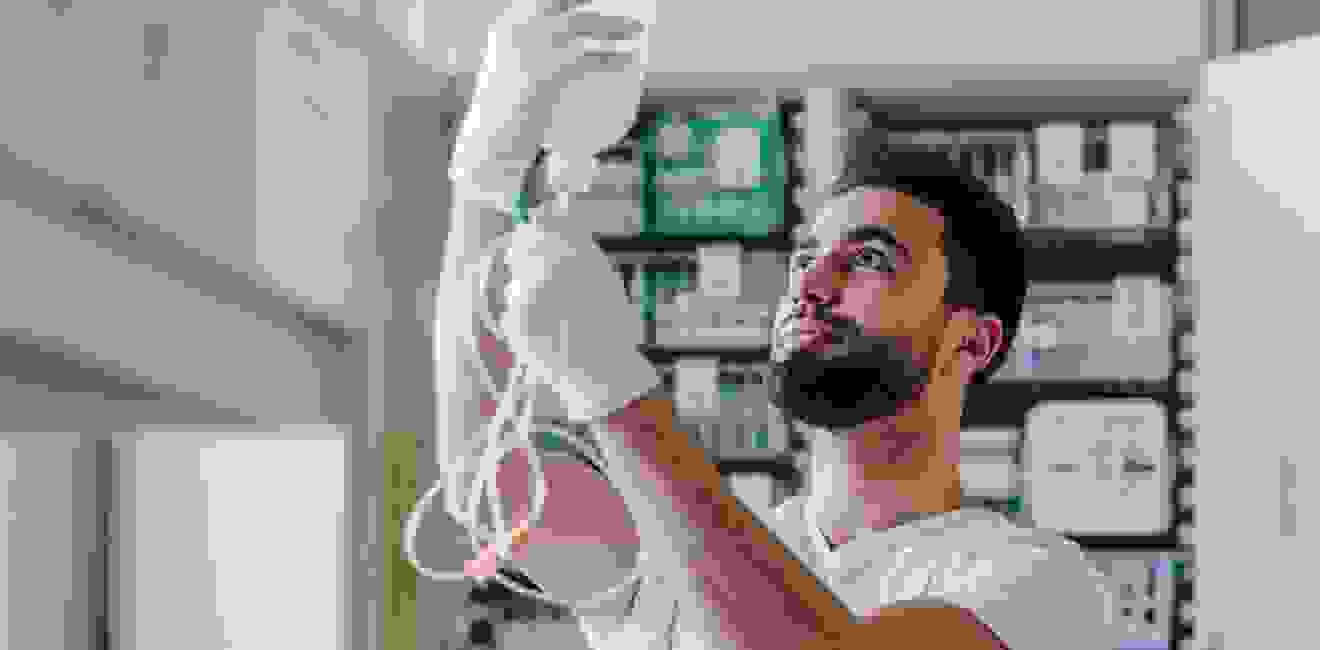 A healthcare worker holds an infusion bottle in his hand and looks at it attentively. Medication and medical equipment are visible in the background.