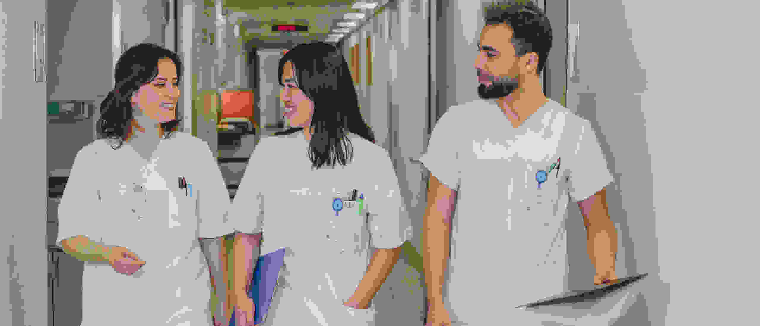 Three nurses in white work uniforms walk down a hospital corridor smiling.