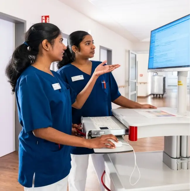 Two nursing staff in blue coats stand at a mobile computer workstation and look at the screen.