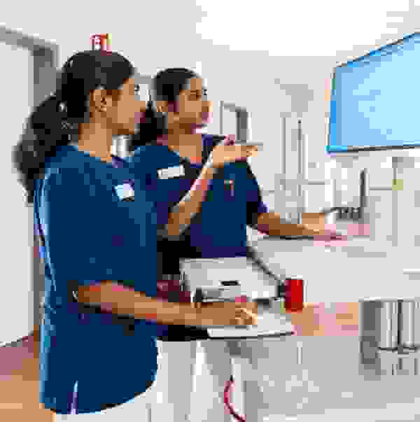 Two nursing staff in blue coats stand at a mobile computer workstation and look at the screen.