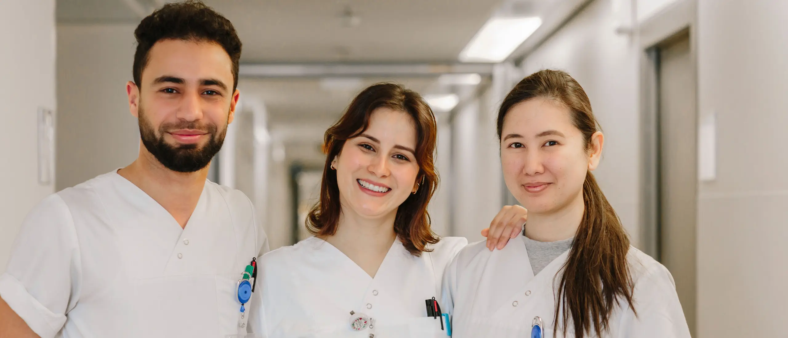 Three cheerful nurses in white work uniforms stand in a hospital corridor and smile at the camera.