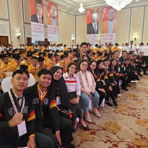 Group photo of participants at an event in a hall where many people wearing orange jackets are sitting. Posters with pictures of leaders are visible in the background.