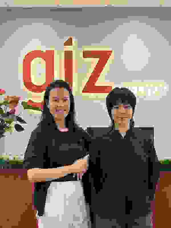 Two women stand in front of a GIZ sign in an office in Hanoi, surrounded by plants and flowers.