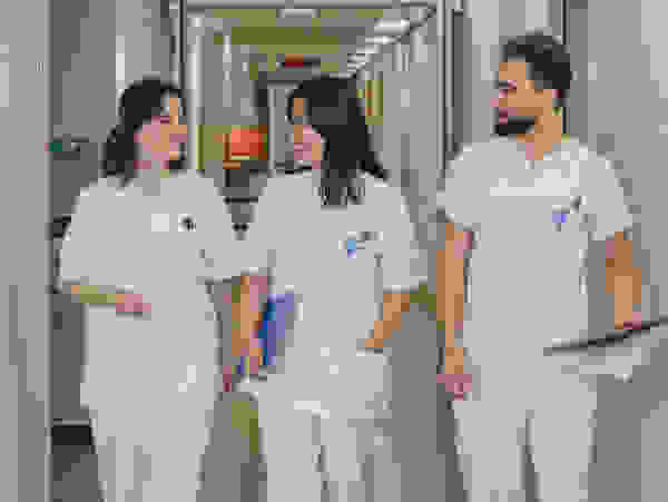 Three medical professionals in white work clothes, smiling as they walk down a hospital corridor, holding notes and documents.