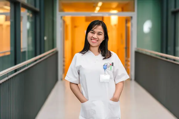Young woman in a white doctor's coat, smiling and standing in a hospital corridor with an orange background.
