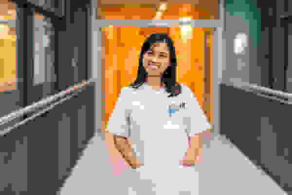 Young woman in a white doctor's coat, smiling and standing in a hospital corridor with an orange background.