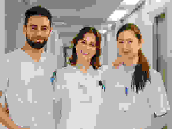 Three cheerful nurses in white work uniforms stand in a hospital corridor and smile at the camera.