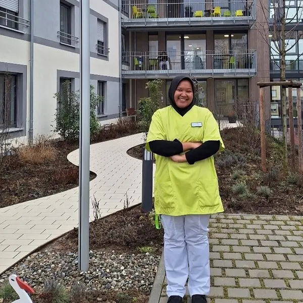 A person in yellow work clothes stands in the inner courtyard of a modern building, surrounded by plants and a sidewalk. Windows with a balcony and a flagpole can be seen in the background.