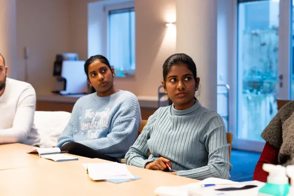 Two women are sitting at a table in a meeting room. One woman is wearing a light blue, structured sweater, the other a blue turtleneck sweater, both are listening attentively. A man can also be seen in the picture, but his focus is not on the camera.