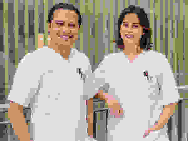 Two smiling healthcare professionals in white coats stand in front of a modern glass wall with vertical stripes.